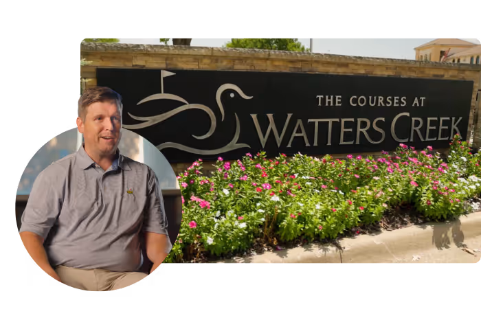 Man in gray polo shirt next to a sign reading The Courses at Watters Creek with flowers in front.
