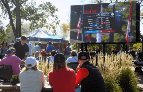 People seated outdoors watching a large screen displaying Ryder Cup golf scores on a sunny day.