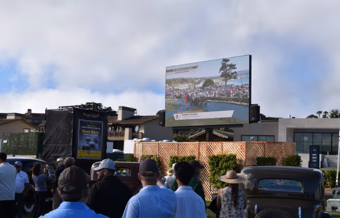 Outdoor crowd watching a large screen showing a golf event with vintage cars parked nearby.