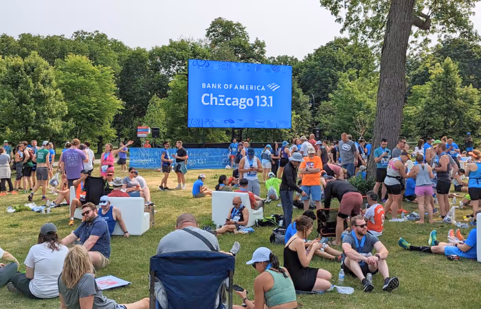 Crowd of runners resting on grass near a large screen displaying Bank of America Chicago 13.1 event.