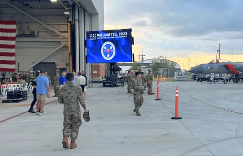 Military personnel stand near a jet on runway with a screen displaying 'William Tell 2023' event logo.