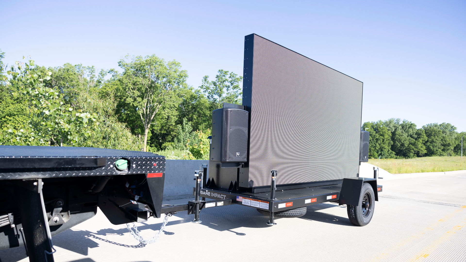 Black mobile LED video screen trailer connected to a truck on a sunny day with trees in the background.