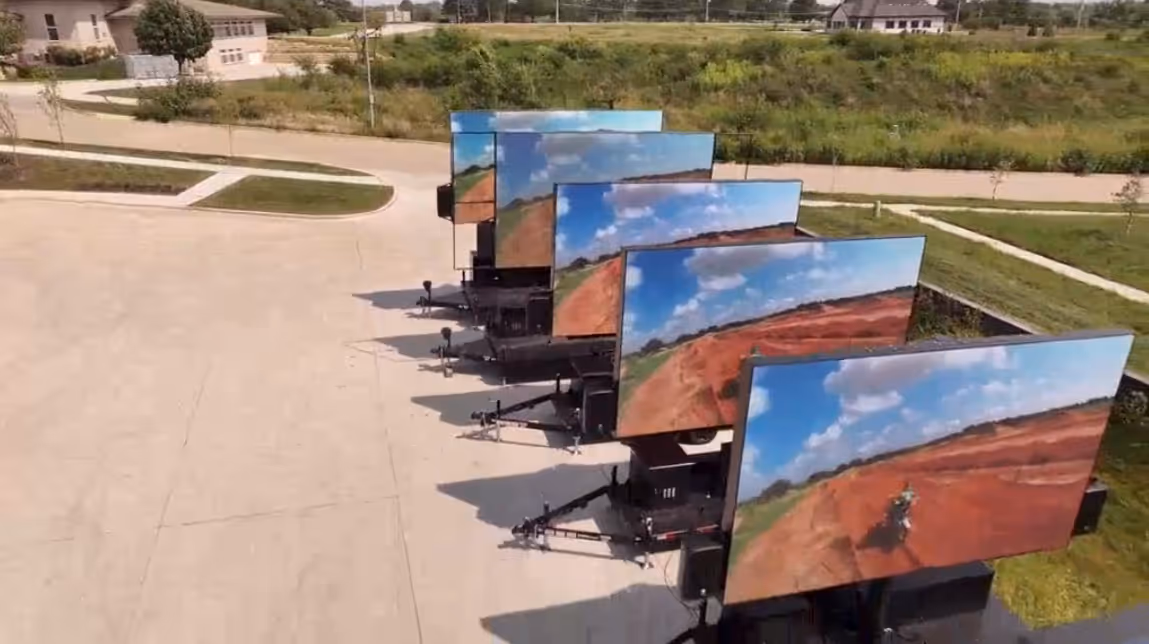 Five large outdoor digital screens in a row showing a dirt bike on a red clay track with blue sky.