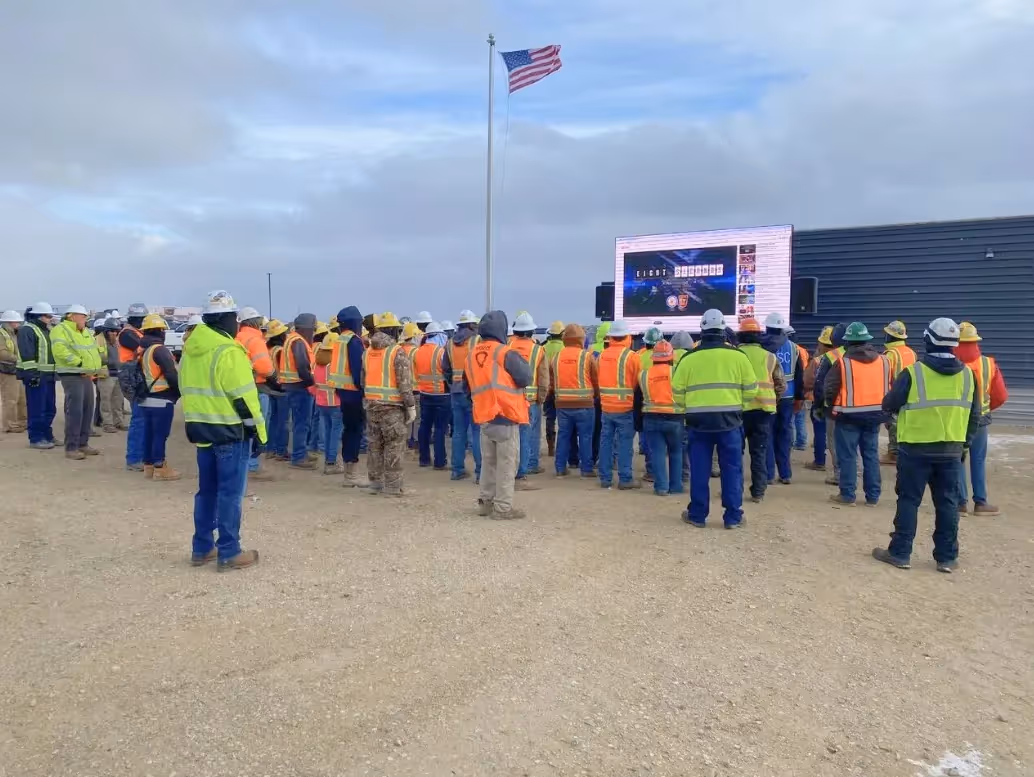 Group of construction workers in high-visibility vests and helmets watching a large outdoor screen with US flag overhead.