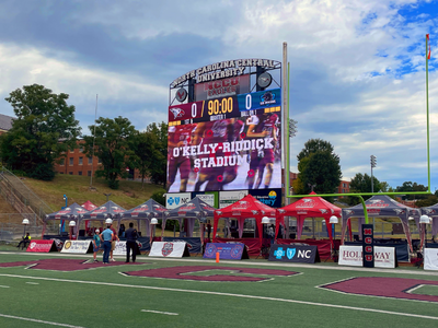 Scoreboard at O’Kelly-Riddick Stadium showing 0-0 score and a row of tents with people on a football field.