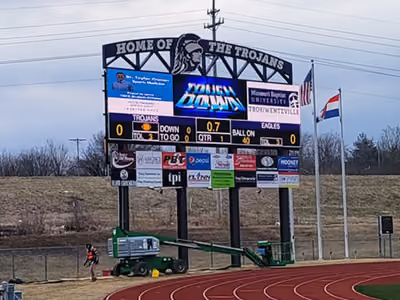 Electronic scoreboard at a track field showing home of the Trojans and current football game stats.