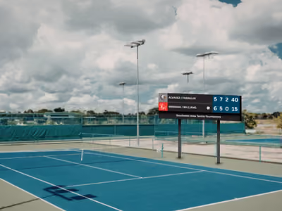 Outdoor tennis court with scoreboard showing match scores under a cloudy sky.