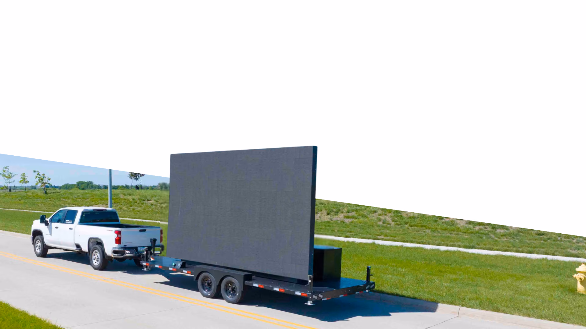 White pickup truck towing a trailer with a large blank LED screen on a road.