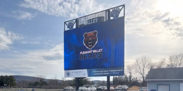 Outdoor scoreboard with Pleasant Valley Bears logo under cloudy sky and parked cars nearby.