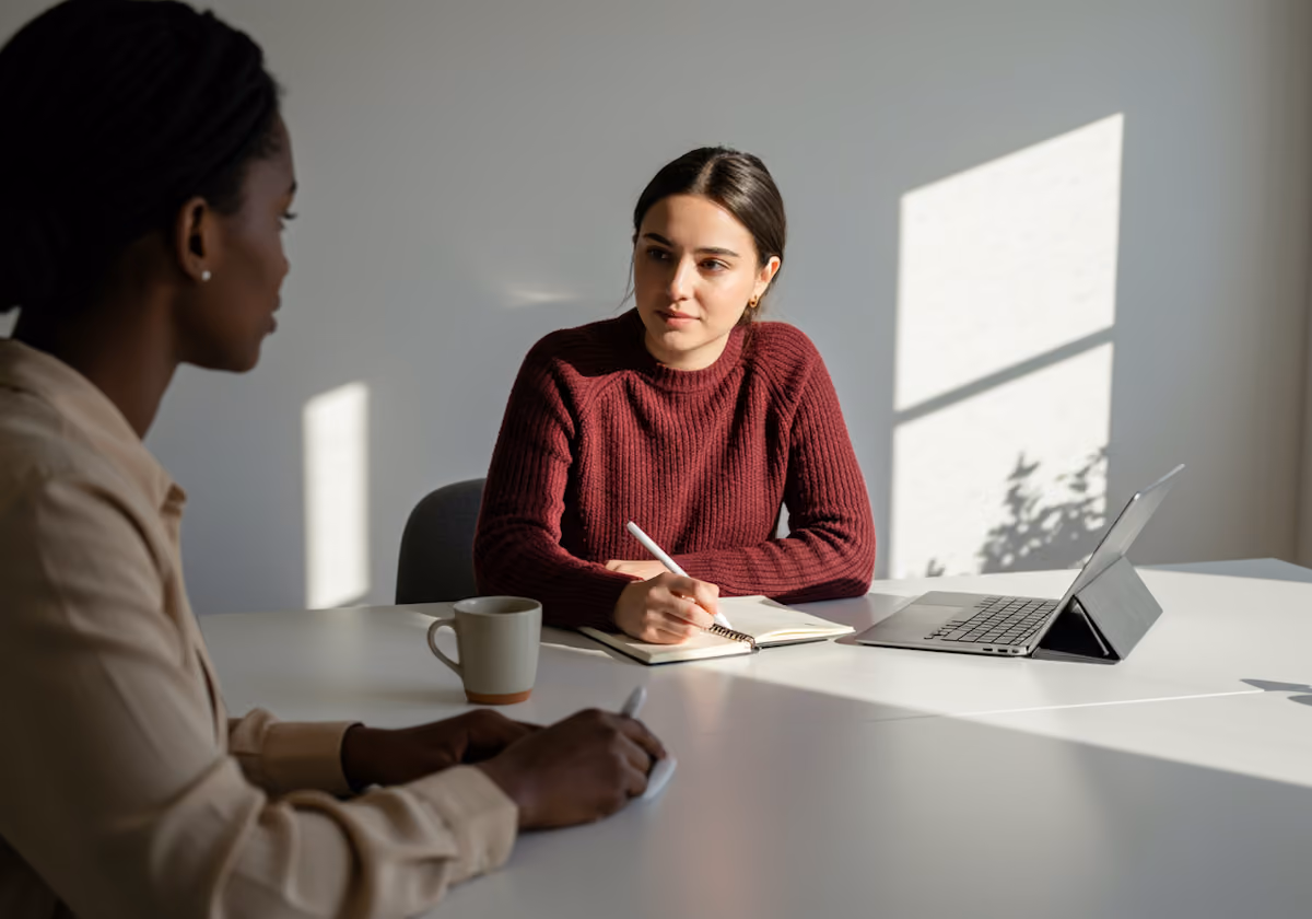 Twee vrouwen zitten aan een tafel in een lichte kamer, één maakt aantekeningen in een notitieboekje terwijl ze luistert, de ander lijkt te spreken.