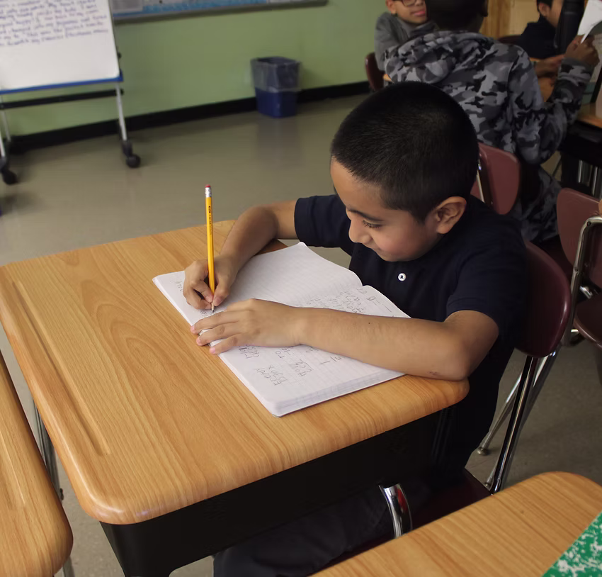 Student writing in notebook with pencil at classroom desk