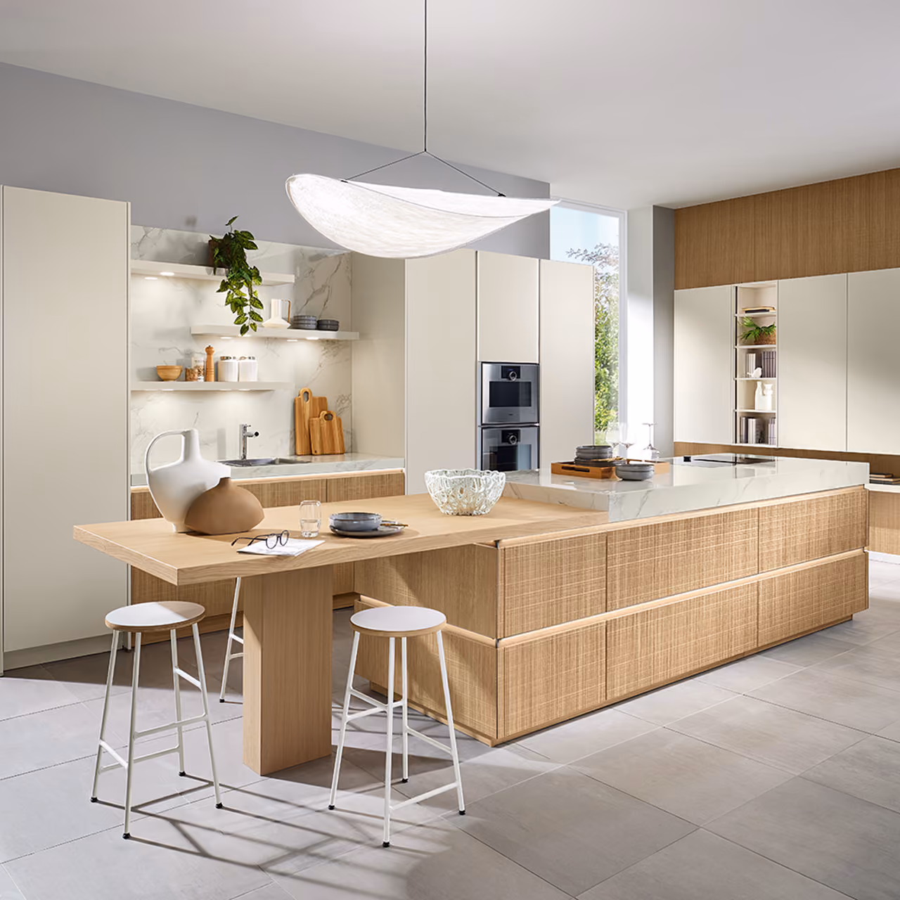 Moderne Küche mit einer großen Kochinsel aus hellem Holz mit weißer Marmorarbeitsplatte, zwei weißen Hockern, Einbaubacköfen und hellen Schränken, einer Hängeleuchte und offenen Regalen mit Küchengeschirr und Grünpflanzen.