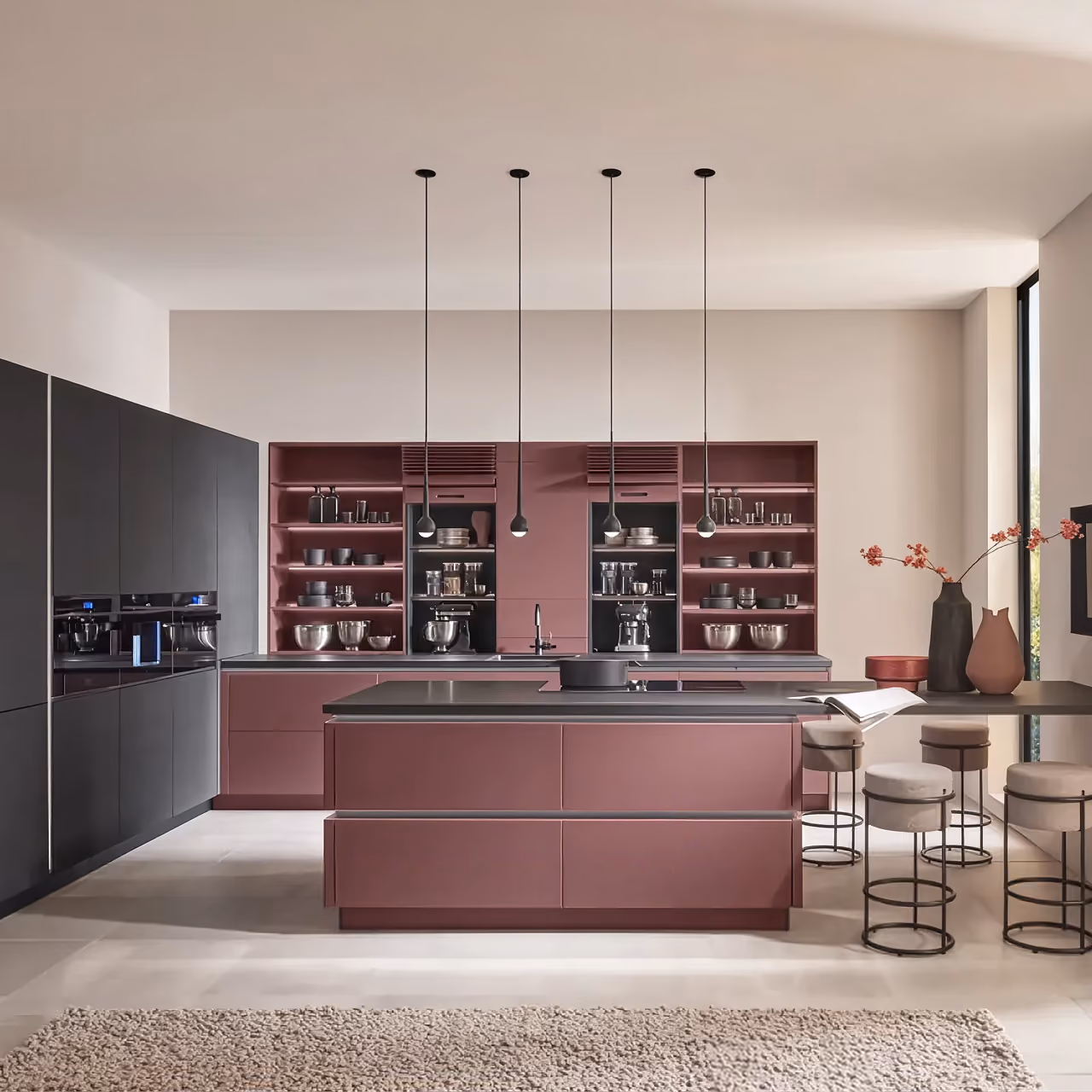 Moderne Küche mit matten weinroten Schränken und Kücheninsel, schwarzen Geräten und vier hängenden Pendelleuchten, daneben hoher Tisch mit vier gepolsterten Barhockern und dekorativen Vasen mit roten Zweigen vor großem Fenster.