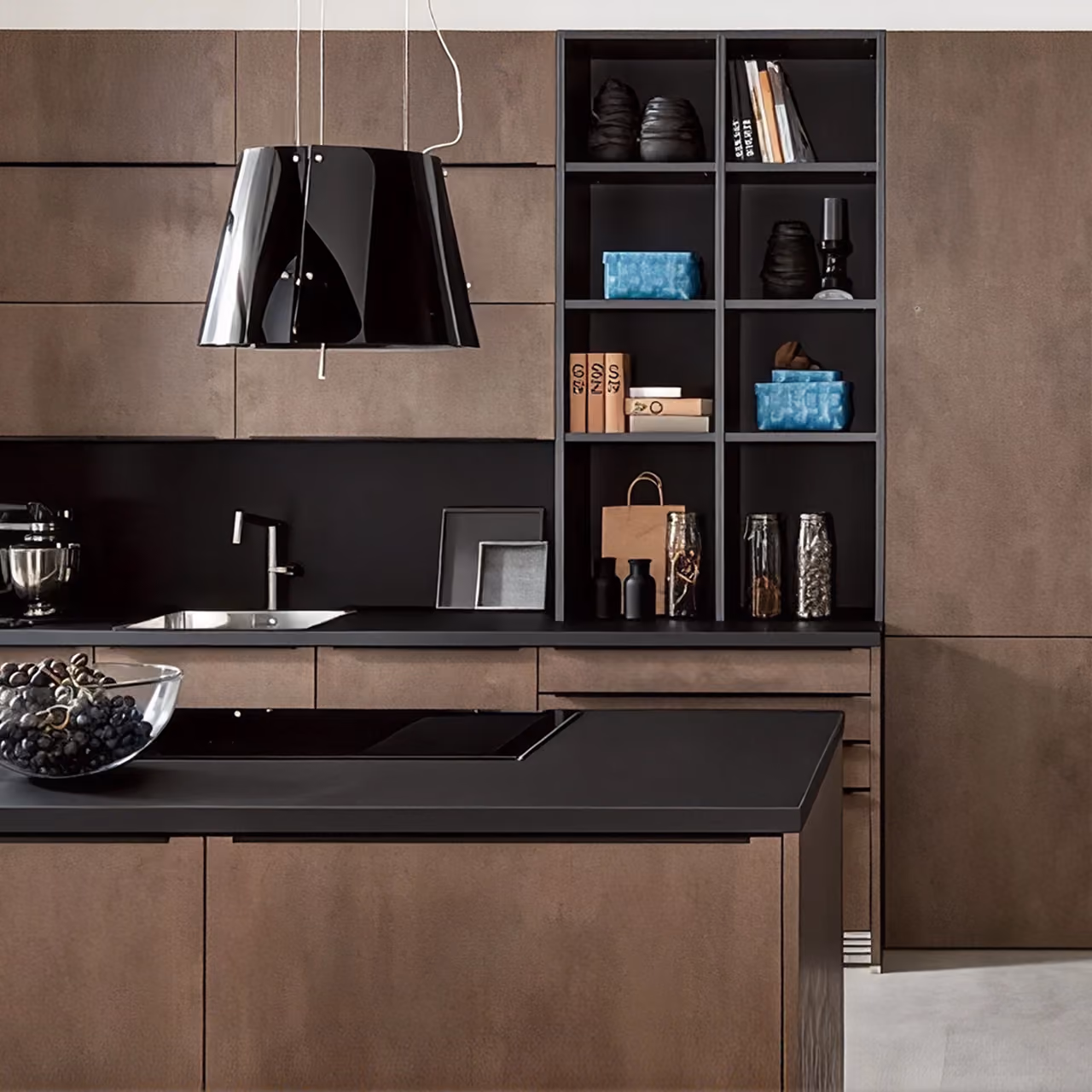 Moderne Küche mit mattschwarzer Arbeitsplatte und braunen Holzschränken; schwarze hängende Lampen, offene Regale mit Dekorationsgegenständen und Kochbüchern, Glas mit Trauben auf der Insel.