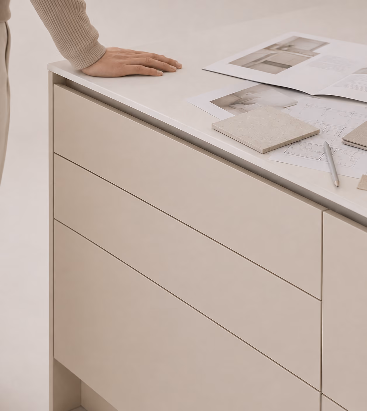 Eine Hand ruht auf einer beigen Küchenarbeitsplatte mit drei Schubladen, daneben liegen Fliesenmuster, ein Stift und ein aufgeschlagenes Magazin mit Küchenfotos.