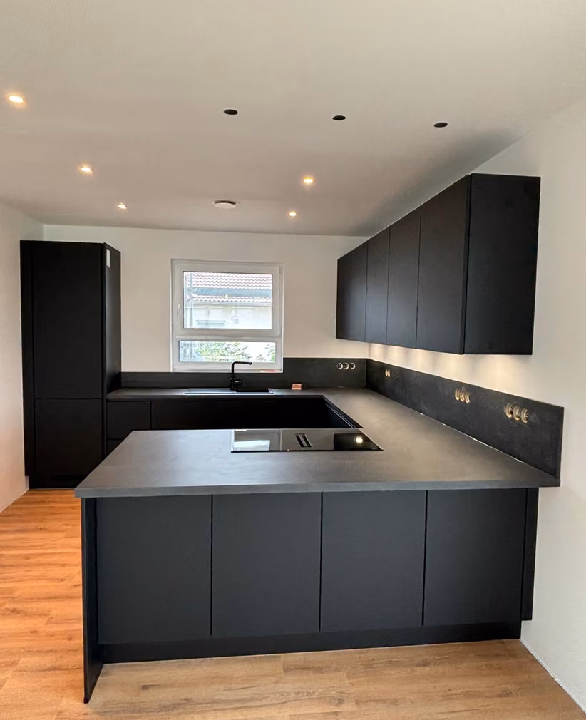 Moderne Küche mit mattschwarzen Schränken und grauer Arbeitsplatte in U-Form, Holzfußboden und einem Fenster über der Spüle.