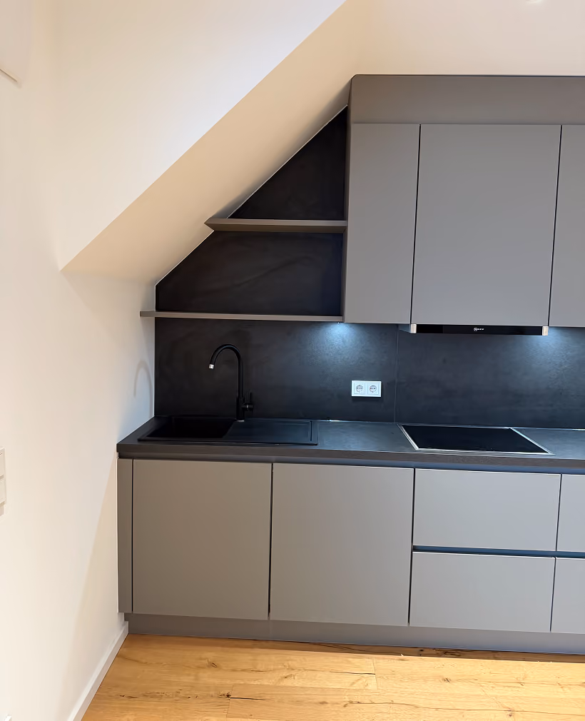 Moderne, minimalistische Küche mit grauen Schränken und Schubladen, schwarzer Spüle und Armatur, schwarzer Arbeitsplatte und Rückwand sowie eingebautem Kochfeld unter einer Dachschräge, heller Holzboden.