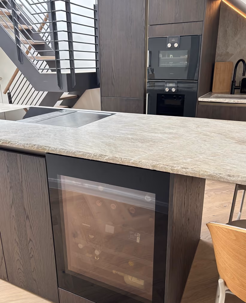 Moderne Küche mit einer Insel aus dunklem Holz und heller Steinplatte, einem eingebauten Weinkühler und einem schwarzen Ofen in der Rückwand, neben einer Treppe mit Metallgeländer.