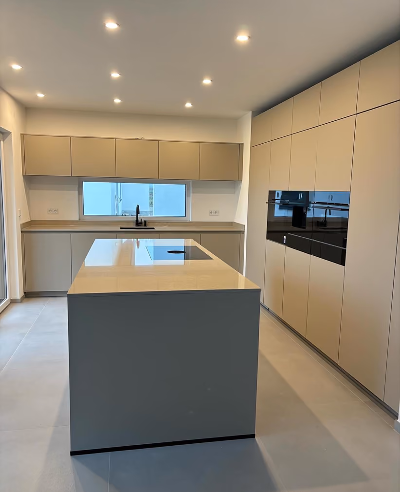 Moderne, minimalistische Küche mit beigen Schränken, Kücheninsel mit Kochfeld, großer Fenster über der Spüle und eingebauten schwarzen Elektrogeräten an der Wand.