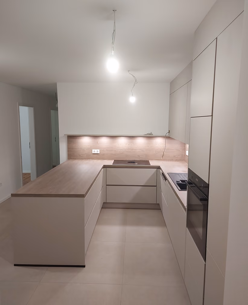 Moderne minimalistische Küche mit hellen Schränken und Arbeitsplatten in Beige- und Holzoptik, eingebautem Backofen und Kochfeld, zwei Deckenlampen und einer Küchenzeile in U-Form.