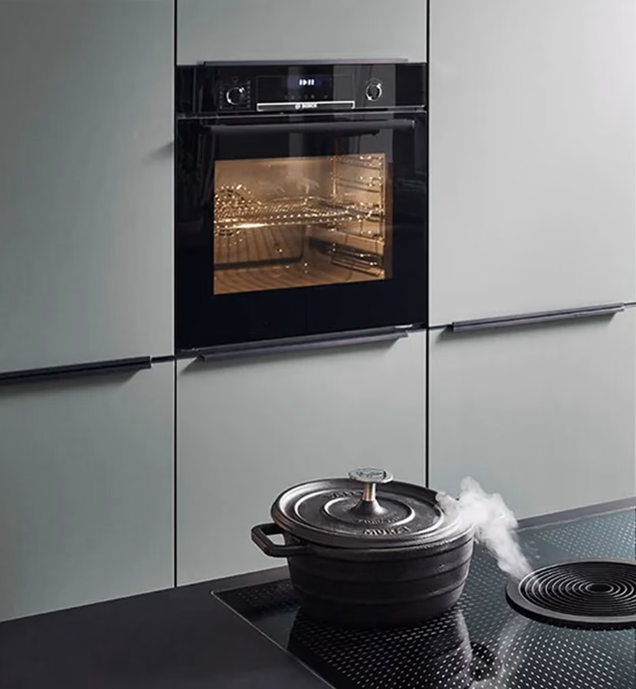 Moderne Küche mit eingebautem schwarzen Backofen in grauen Schränken und einem schwarzen gusseisernen Topf mit Deckel, aus dem Dampf auf einer schwarzen Herdplatte aufsteigt.
