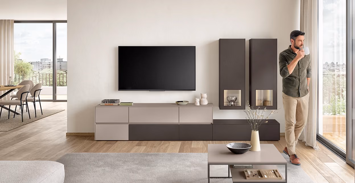 Moderner Wohnraum mit hellem Holzboden, einer grauen Wohnwand mit eingelassenem TV, zwei hohen Wandregalen, einem Couchtisch mit Dekoration und einem Mann, der aus einem Becher am großen Fenster blickt.
