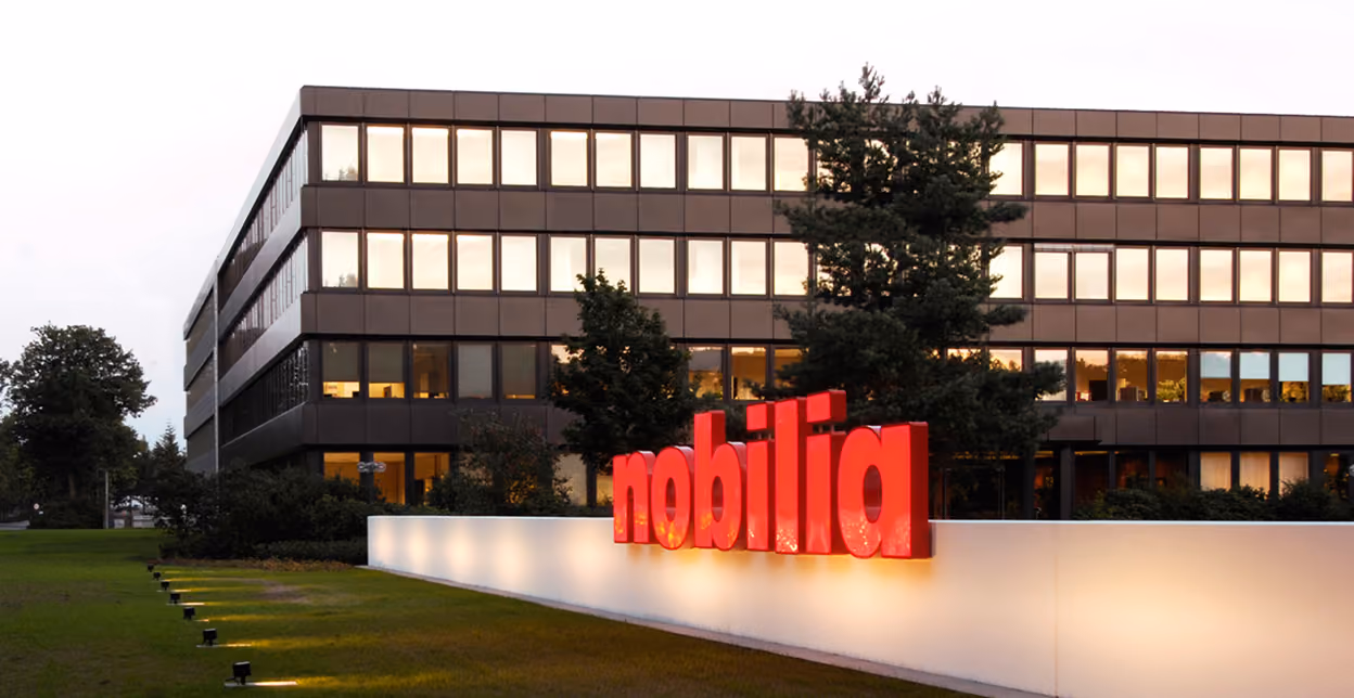 Firmengebäude mit mehreren Fenstern bei Dämmerung, im Vordergrund leuchtendes rotes Schild mit der Aufschrift "nobilia" vor einer weißen Wand mit Beleuchtung.