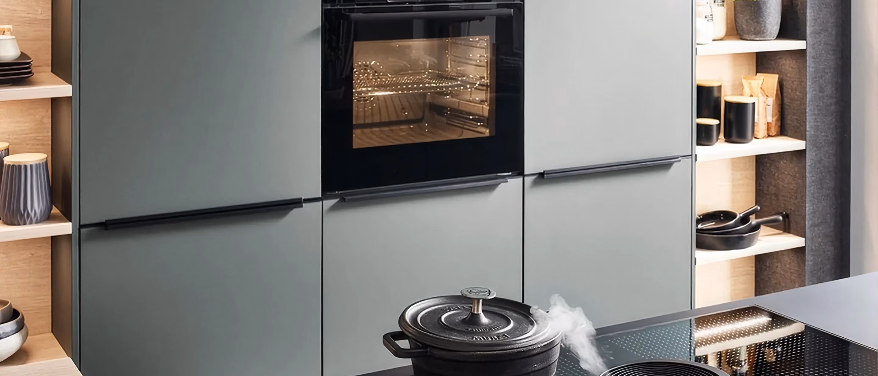 Moderne Küche mit grauen Einbauschränken, einem schwarzen Elektrobackofen in der Mitte und einem Dampfkochtopf auf dem Induktionskochfeld vorne rechts.
