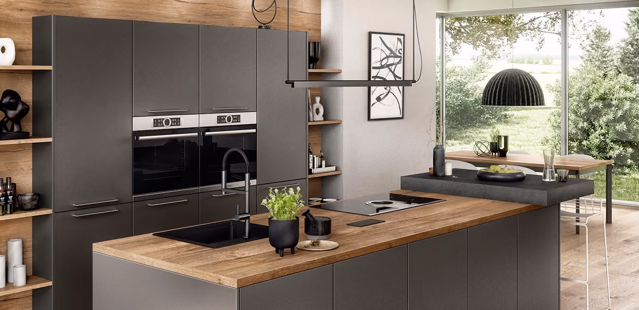 Moderne Küche mit mattschwarzen Schränken und Holzarbeitsplatten, einem eingebauten Ofen, einem schwarzen Spülbecken mit Wasserhahn, offener Regale mit Dekoration, großen Fenstern mit Blick ins Grüne und einem schwarzen Hängelampe über einer Kochinsel mit Sitzplätzen.