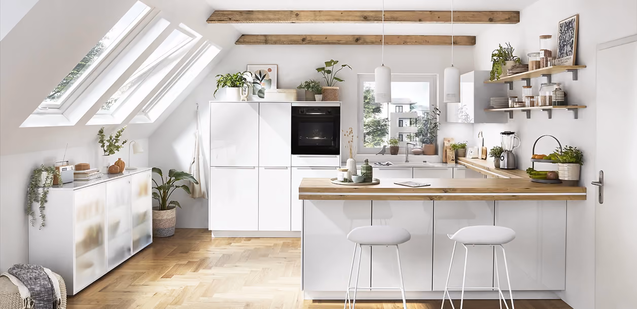 Moderne weiße Küche mit Holzakzenten, zwei Hockern an der Insel, helles Parkett, Fenster im schräge Dach, und Pflanzen auf Regalen und Arbeitsflächen.