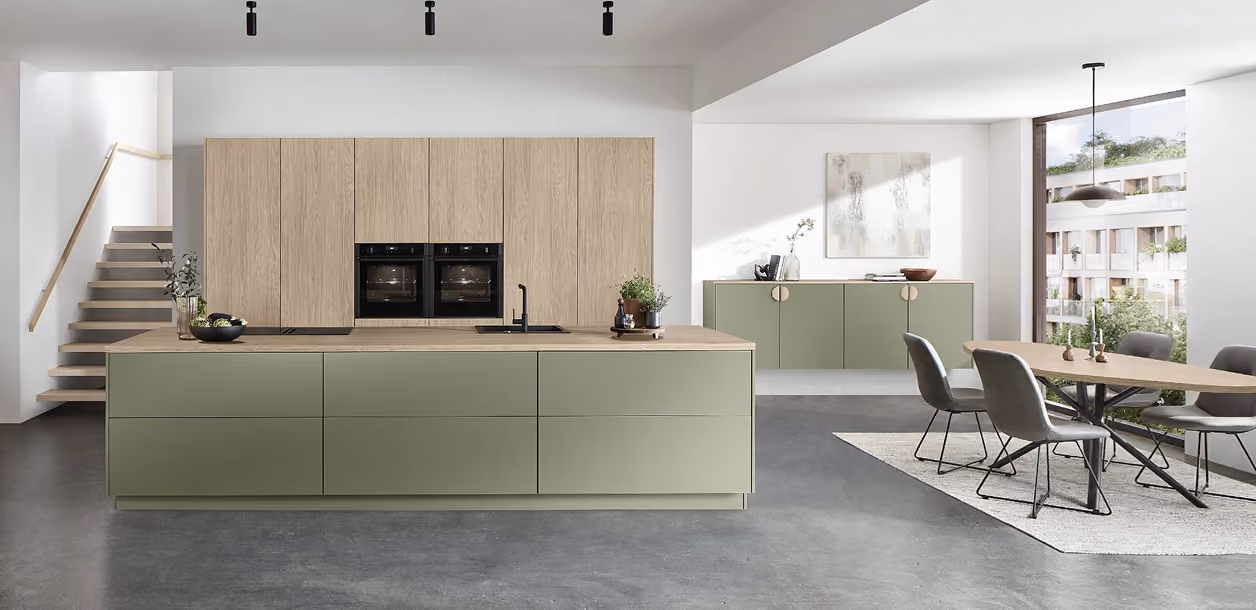 Moderne Küche mit Kücheninsel in mattem Grün und Holzfronten, integriertem Backofen, Kochfeld und Spüle, angrenzender Essbereich mit Esstisch, vier grauen Stühlen und großem Fenster mit Blick auf Wohngebäude.