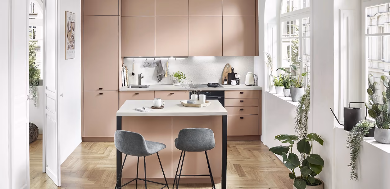 Moderne Küche mit matten rosafarbenen Oberschränken und Unterschränken, mittig steht eine Kücheninsel mit weißer Arbeitsplatte und zwei grauen Barhockern, große Fenster an der rechten Seite mit mehreren grünen Pflanzen auf der Fensterbank und hölzernen Parkettboden.