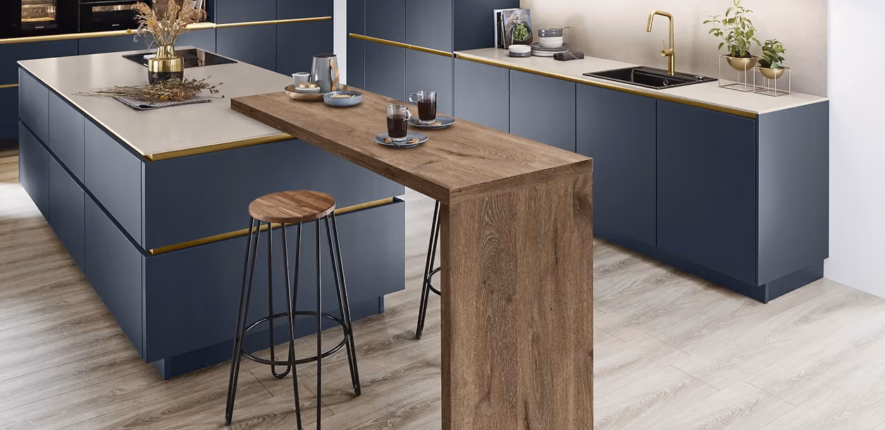 Moderne Küche mit dunkelblauen Schränken, goldfarbenen Griffleisten, einer Kücheninsel mit weißer Arbeitsplatte und einem höhenverstellbaren Bartisch aus Holz mit zwei Barhockern, heller Holzoptik-Bodenbelag und minimalistischer Dekoration.