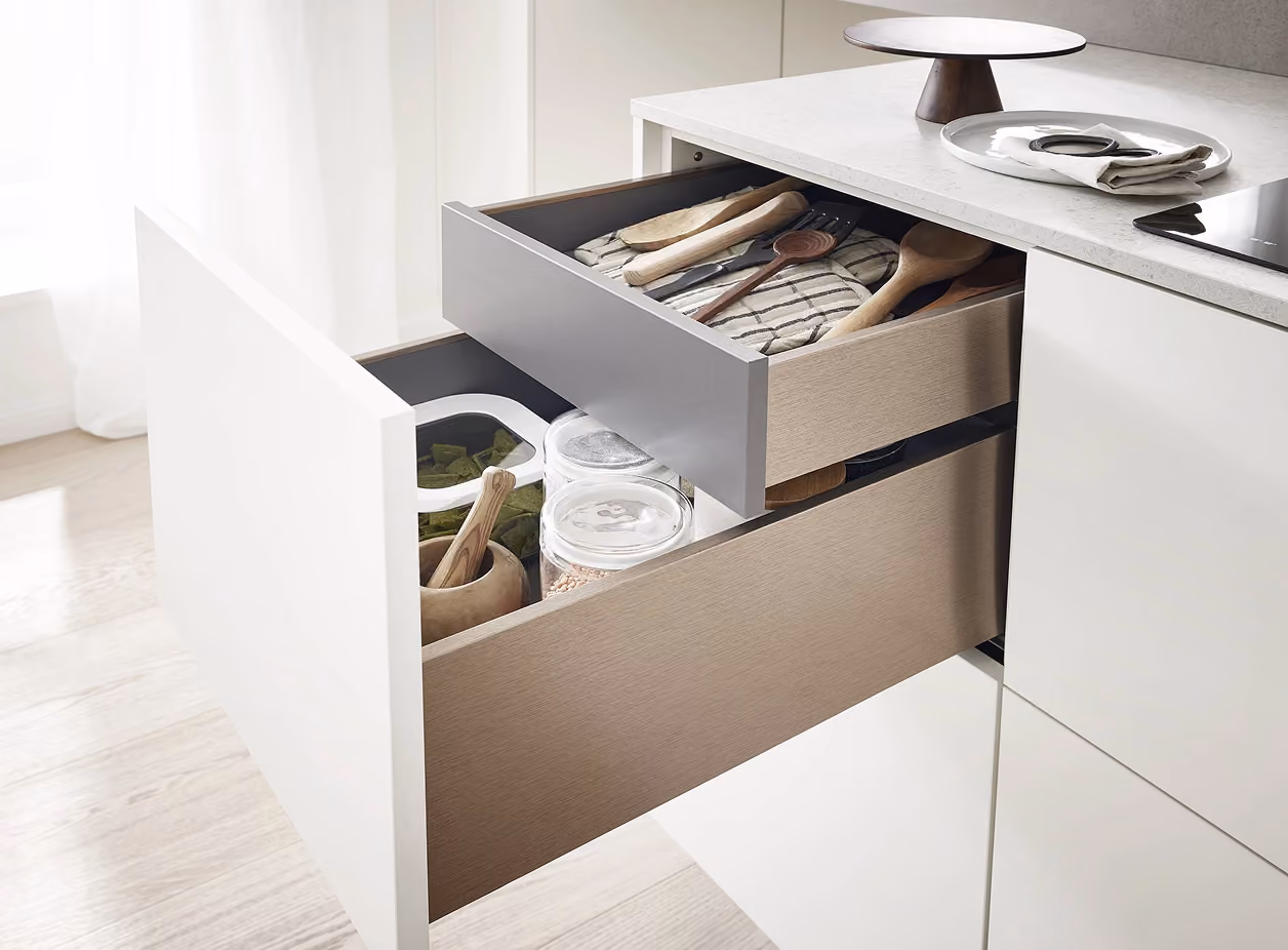 Offene Küchenschubladen mit modernen Holzlöffeln, Küchenutensilien, Gewürzgläsern und einem Mörser, in einer hellen, minimalistischen Küche mit weißer Arbeitsplatte und Holzfußboden.