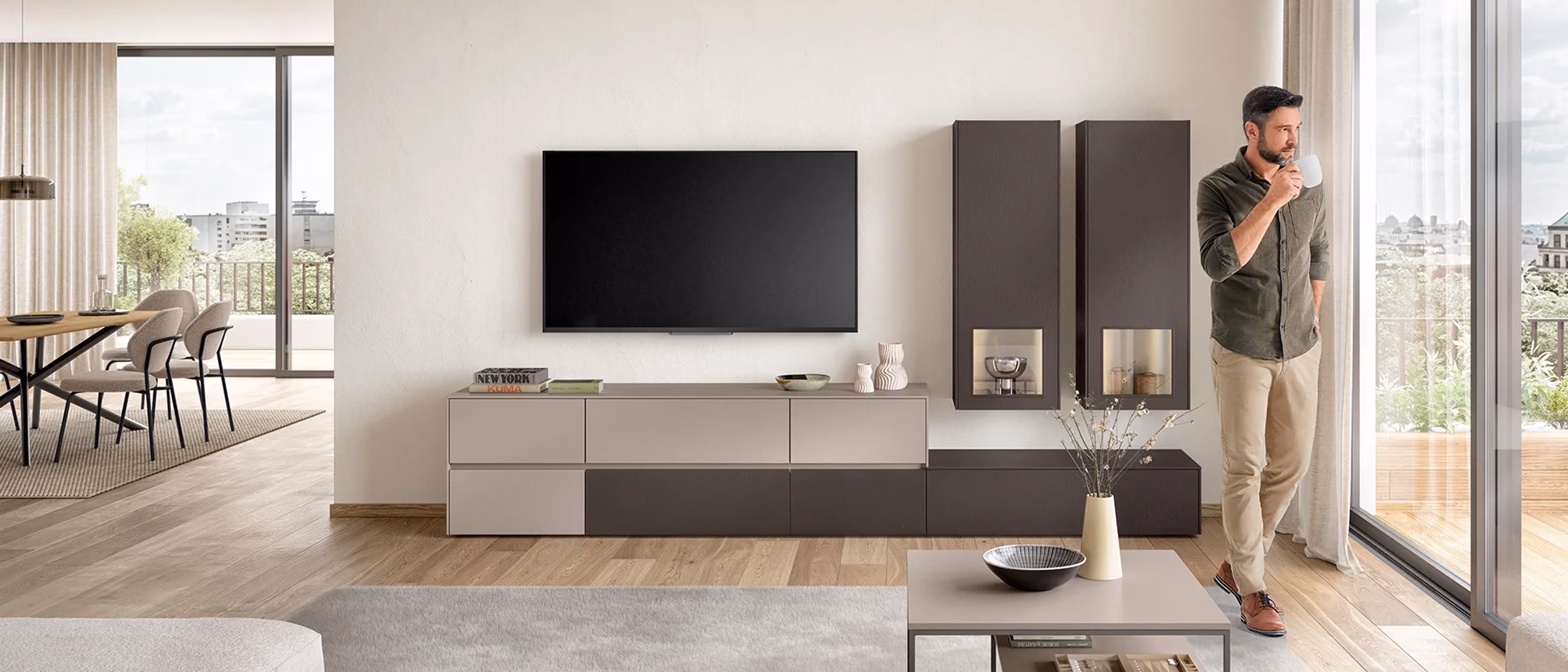 Moderner Wohnbereich mit hellem Holzfußboden, grauem Sitzbereich und passend kombiniertem Sideboard unter einem an der Wand montierten Fernseher, rechts steht ein Mann mit einer Tasse vor einer großen Fensterfront mit Blick auf eine Terrasse und Stadt.