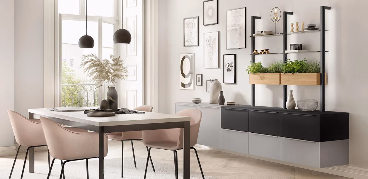 Modern dining area with a light gray table and four pink upholstered chairs with black legs, next to a large window. The wall features framed minimalist artwork, and a sleek black and gray wall-mounted cabinet with wooden planters holding green herbs and various decorative items on shelves.