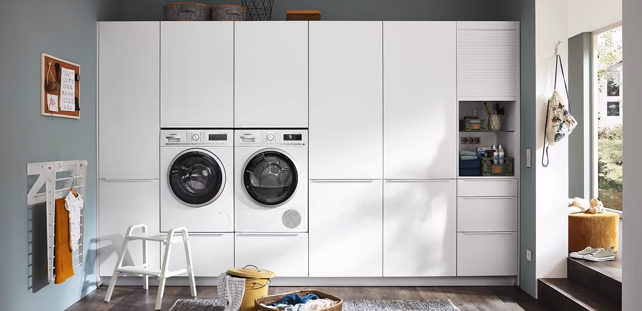 Moderner Hauswirtschaftsraum mit weißen Hochglanzschränken und integrierter Waschmaschine und Trockner, Boden mit Holzoptik und Tageslicht durch Fenster.