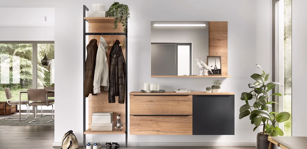 Moderner Eingangsbereich mit einer Garderobe aus Holz und Metall, bestehend aus einer offenen Kleiderstange mit Jacken und einem Regal für Schuhe, daneben eine hängende Kommode mit Schubladen und schwarzer Front, darüber ein beleuchteter Spiegel, rechts eine grüne Pflanze vor einem großen Fenster.