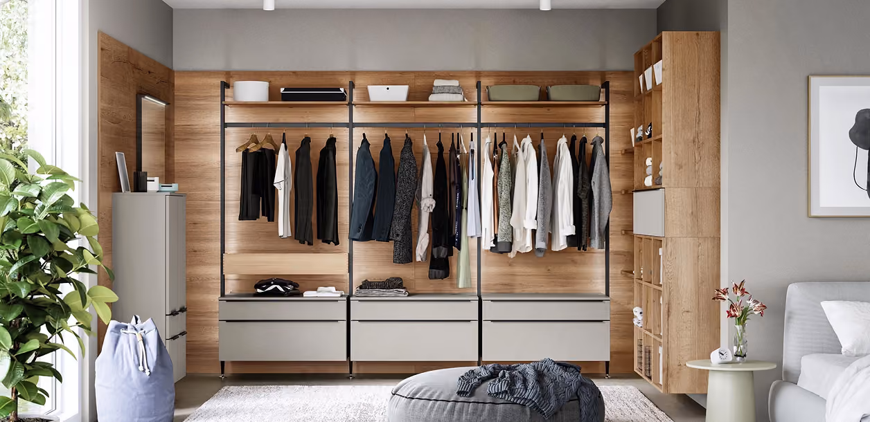 Moderne Ankleide mit offenen Holzregalen und grauen Schubladen, auf denen Kleider und Jacken hängen, daneben ein Spiegel, Pflanzen links und ein gemütliches Wohnzimmer mit Sofa und Bild an der Wand rechts.