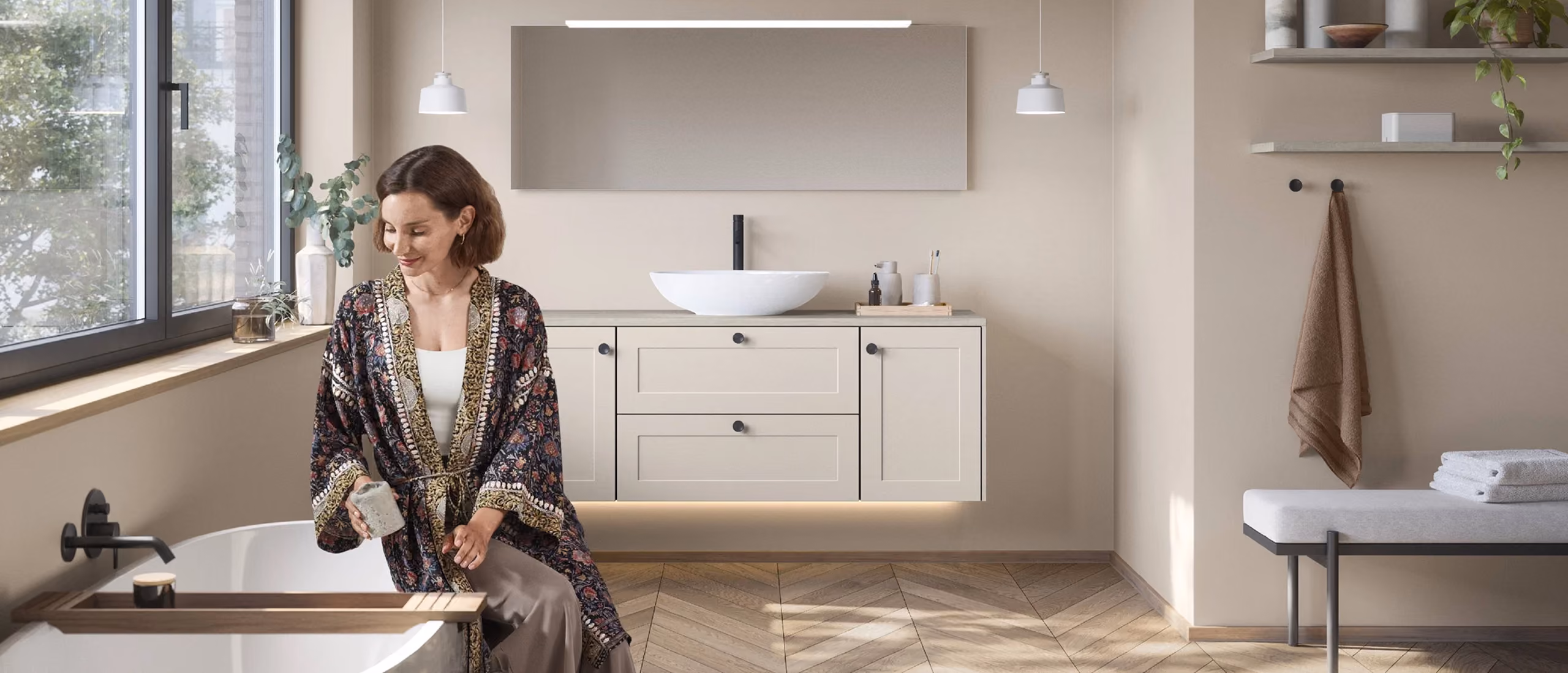 Frau in einem Blumen-Morgenmantel sitzt neben einer freistehenden Badewanne in einem modernen Badezimmer mit hellbeigen Wänden, einem wandmontierten Waschtisch mit rundem Becken und einem rechteckigen Spiegel darüber.