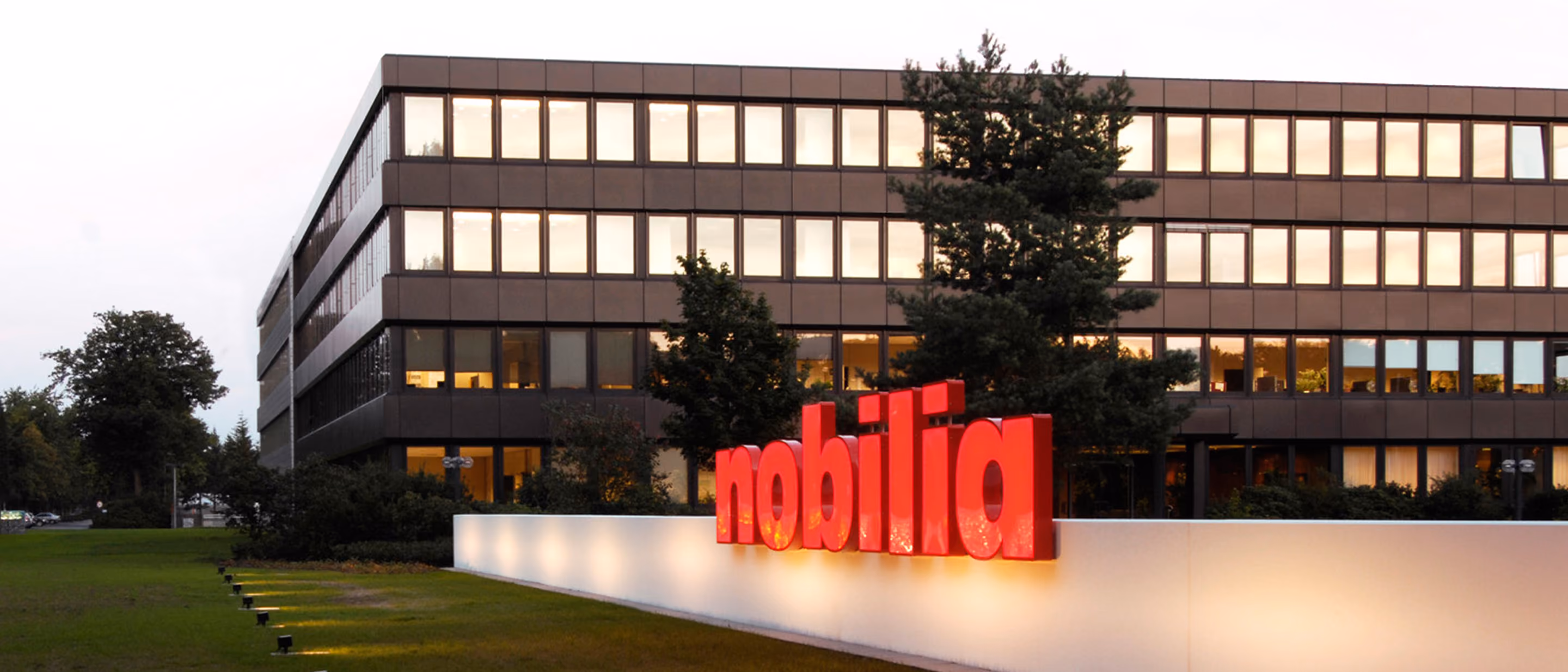 Bürogebäude mit mehreren Fensterreihen bei Abendlicht, davor ein beleuchtetes rotes nobilia-Logo auf einer weißen Mauer, umgeben von Bäumen und Grünfläche.