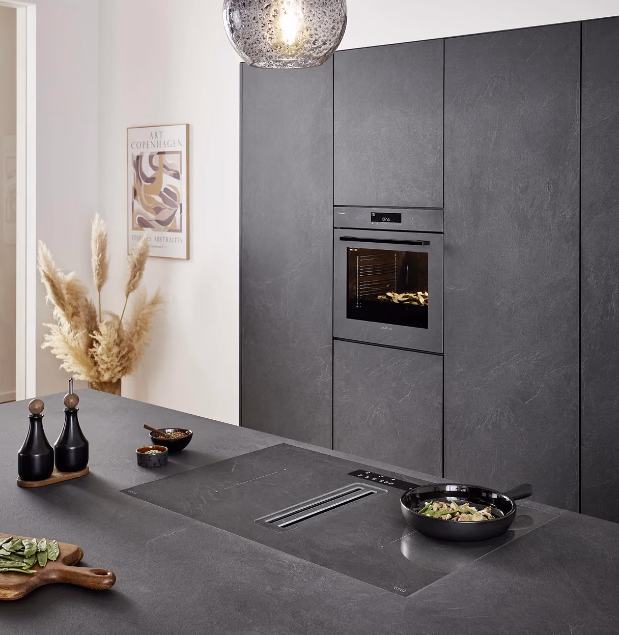 Moderne Küche mit grauer Arbeitsplatte und Einbaugeräten, darunter ein Ofen mit eingeschaltetem Backofen, darüber eine runde Hängeleuchte, links Deko mit getrocknetem Pampasgras und einer abstrakten wandhängenden Kunstdruck.