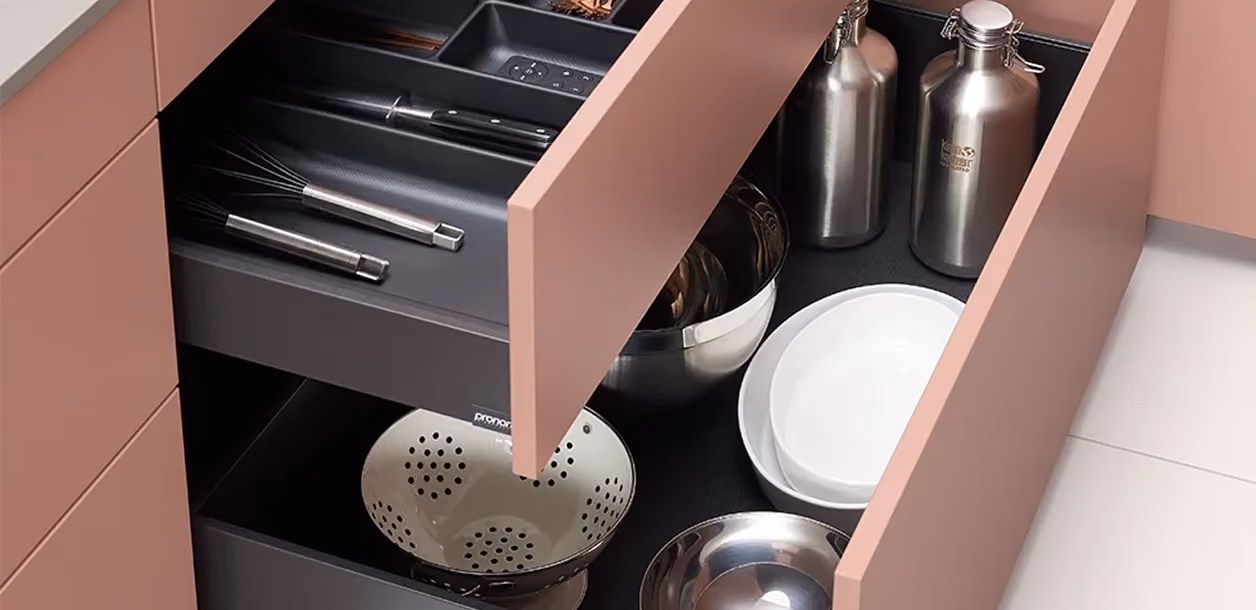 Offene Küchenschubladen mit rosa Fronten zeigen gut organisierte schwarze Einsätze, die Küchenutensilien wie Schneebesen, Schüsseln, Teller und Metallflaschen aufbewahren.