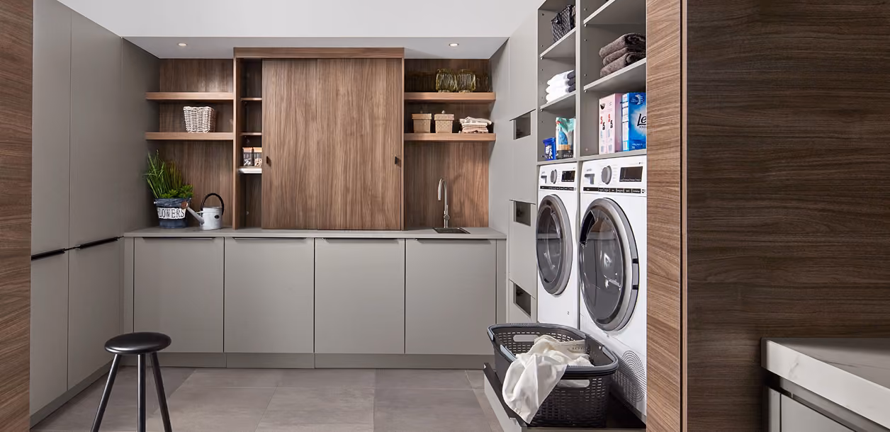 Moderner Hauswirtschaftsraum mit weißen Frontladern für Waschmaschine und Trockner, grauen Schränken und Holzregalen mit Körben und Dekoration, grauem Hocker und einem Wäschekorb mit Wäsche.