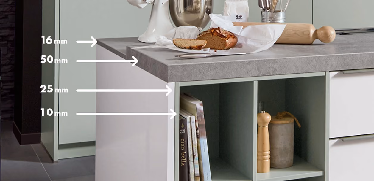 Kücheninsel mit mehrschichtiger Arbeitsplatte, auf der verschiedene Schichtdicken mit weißen Pfeilen und Maßen von 16 mm, 50 mm, 25 mm und 10 mm an der Platte markiert sind, hinten liegen Brot, eine Küchenmaschine und ein Nudelholz.