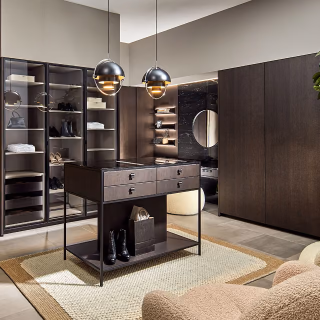 Moderner begehbarer Kleiderschrank mit dunklen Holzschränken, beleuchteten Glasvitrinen, einer Insel mit Schubladen in der Mitte und zwei hängenden schwarzen Pendelleuchten über einem Teppich.