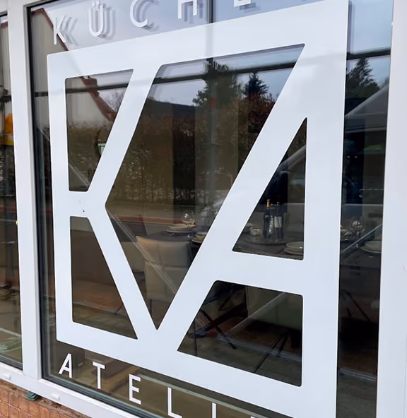 Fenster mit weißem Logo, das stilisiert die Buchstaben K und A zeigt, darüber steht Kü chen und darunter Atelier, mit Spiegelung eines gedeckten Tisches im Inneren und Bäumen draußen.