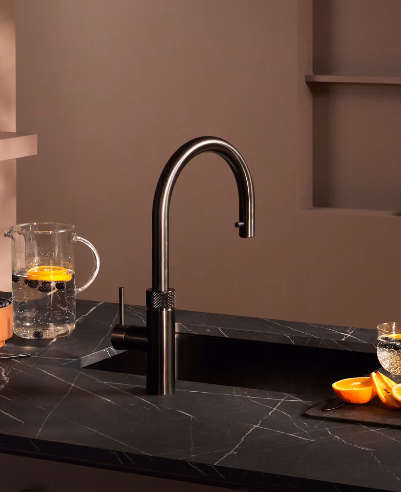 Moderner schwarzer Küchenwasserhahn mit hochwertiger Armatur auf einer eleganten, dunklen Marmorarbeitsplatte, daneben Karaffe mit Wasser, Zitronenscheiben und Beeren sowie ein Glas mit kohlensäurehaltigem Wasser und Orangenscheiben.