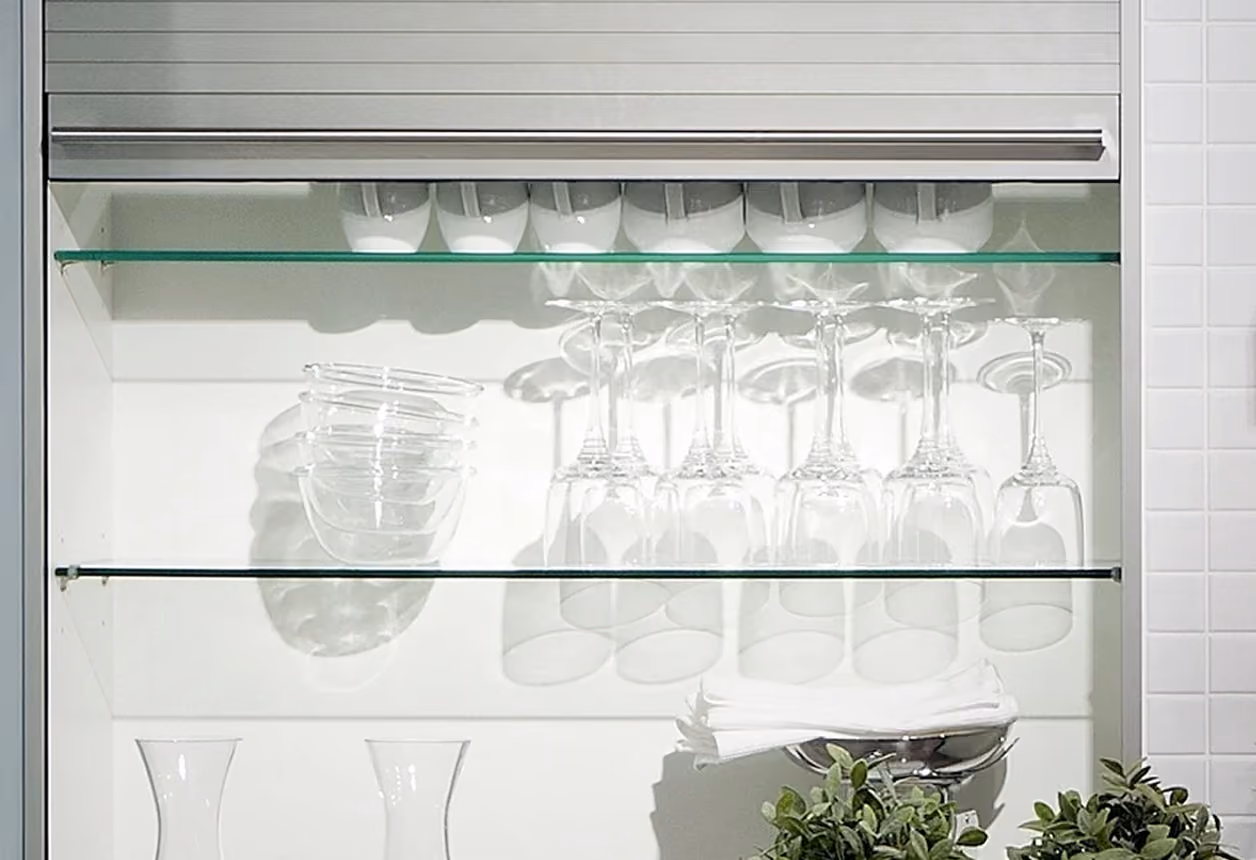 Geschlossener Schrank mit transparentem Rollladenschrank in einer Küche, mit Glasregalen, die Gläser, weiße Schalen und Stoffservietten enthalten.