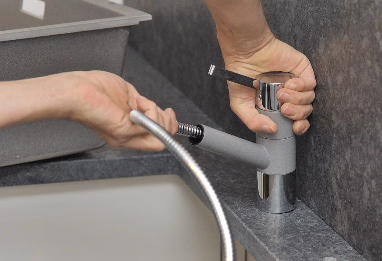 Hand zieht die ausziehbare Brause eines Küchenwasserhahns heraus, die auf einer grauen Arbeitsplatte neben einem Spülbecken montiert ist.